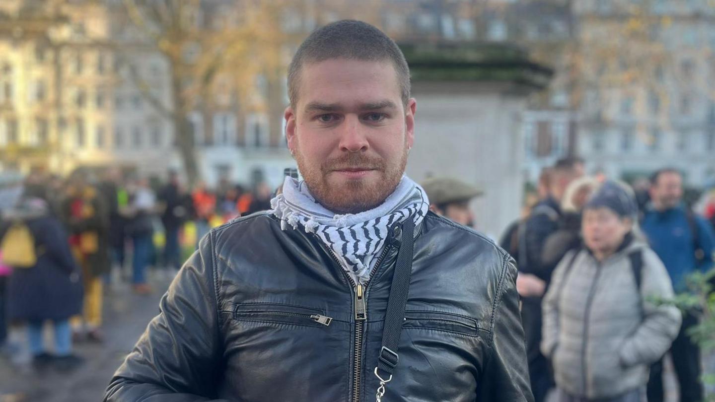 Conall Ó Brolcháin is seen in at a demonstration wearing a leather jacket and a scarf. 