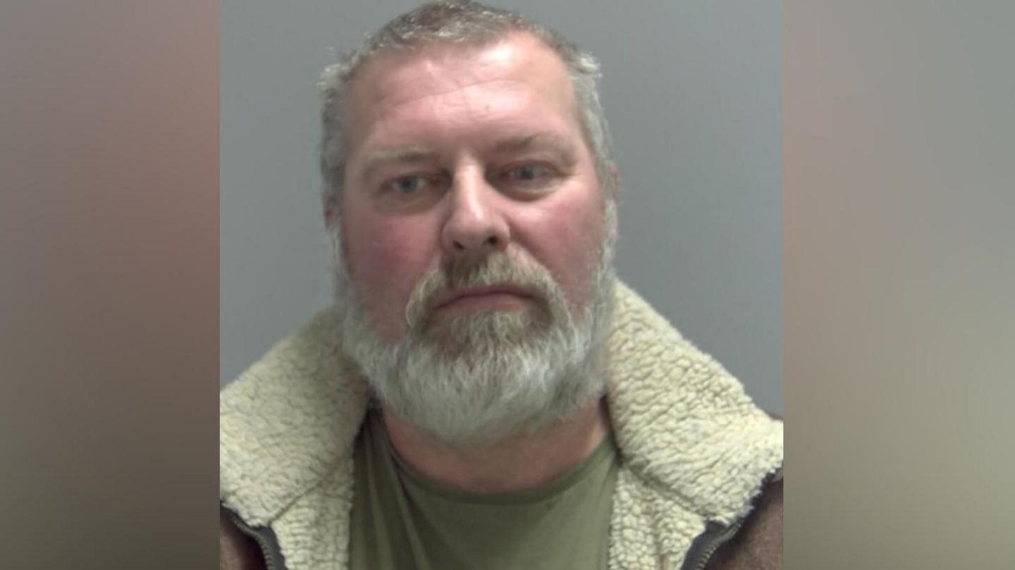 Trevor New-Watts is looking at the camera in a police custody shot. He has short greying hair and facial hair. He is wearing a green top and a brown jacket with a cream collar.