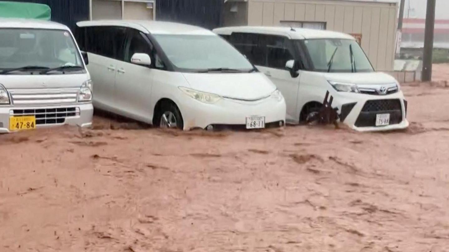 Brown floodwater gushes down a road in central Japan. Three white vehicles are parked with the water up to its wheels and bumpers