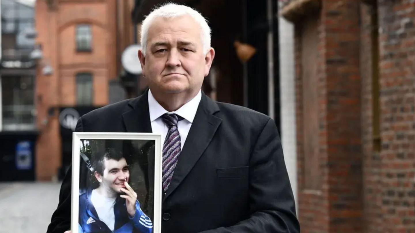 Mr Brown has short grey hair and is wearing a black suit jacket, striped tie and white shirt as he holds an image of his son Aaron.