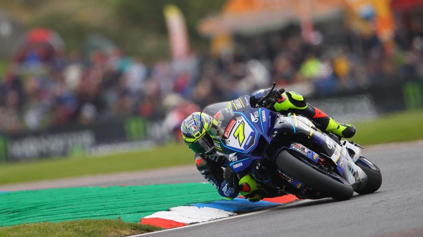 Ryan Vickers in multi-coloured OMG Racing Yamaha gear and helmet, taking a tight turn on a bend on his bike, in front of an onlooking crowd at a British Superbike meeting.