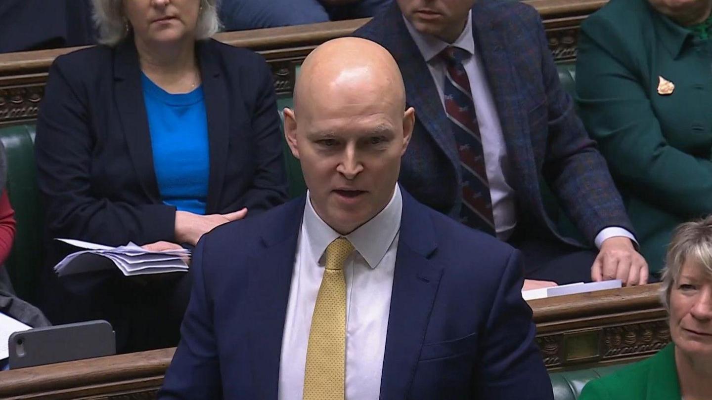 A screenshot of Max Wilkinson talking in Parliament this morning. He is bald with very fair eyebrows, and wears a dark suit with a white shirt and yellow tie. Other MPs can be seen behind him on the green leather benches. 