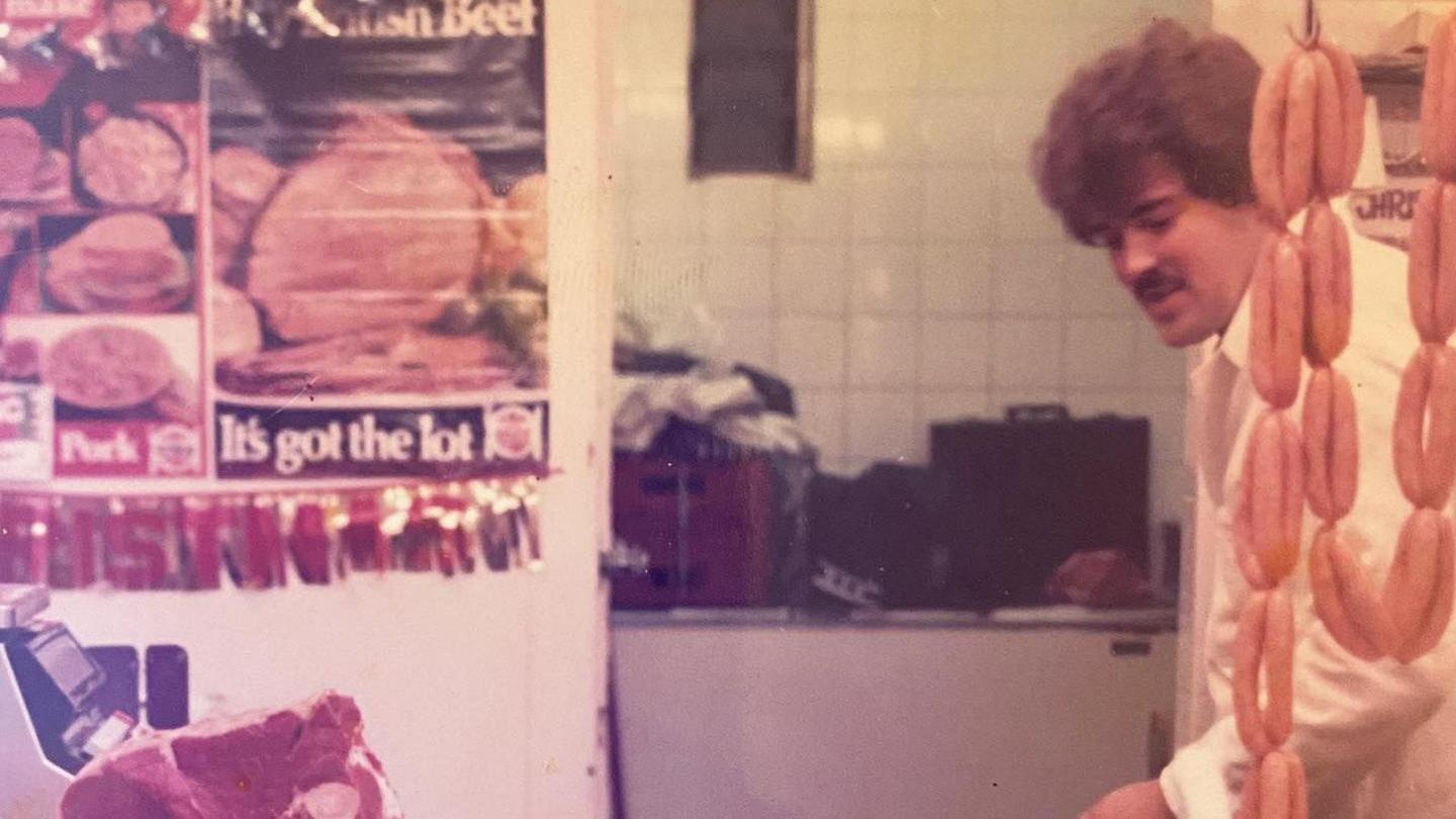 An older photos of Geoff Hughes. who has thick brown hair and a moustache, and is peeping out from behind a string of sausages.