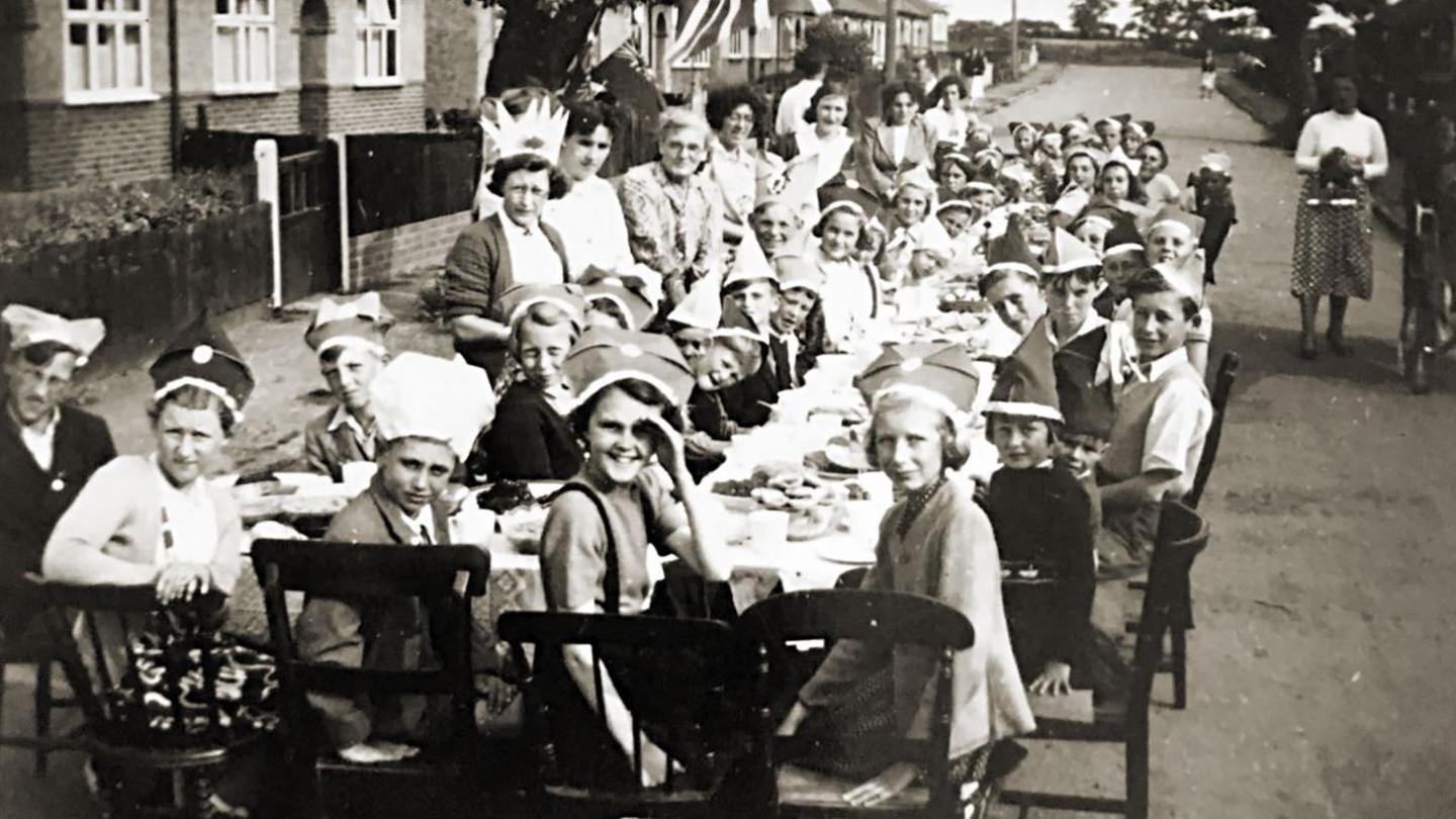 A black and white photo of a group of children and adults enjoying a street party, with food being shared on a long table in the middle of a residential street. 