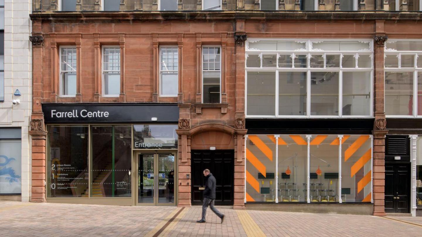 The Farrell Centre is part of a row of buildings. It is made of red stone with tall glass front windows. An exhibition space in the building to the left has an orange stripey design in the window.