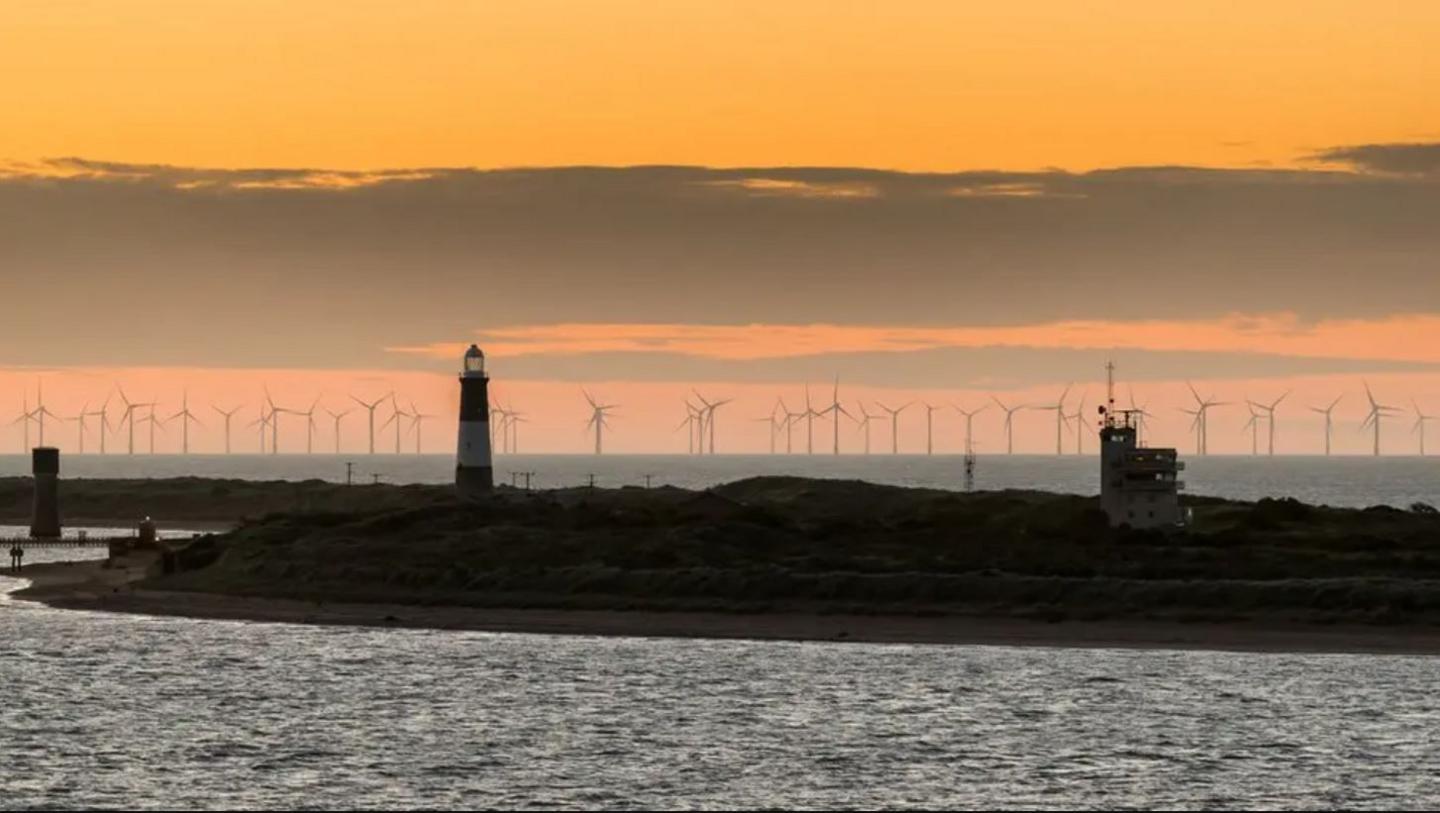A view of a thin spit of land sandwiched between the North Sea and the Humber Estuary featuring a lighthouse. Multiple wind turbines can be seen in the distance. The sky is orange and cloudy.