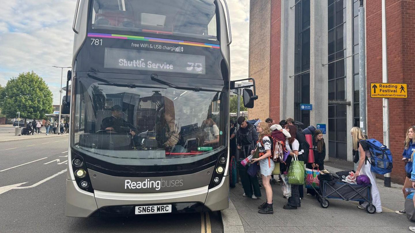 A group of festivalgoers with bags board a Reading Buses shuttle labelled "Shuttle Service" on a city street, with a sign for Reading Festival nearby.
