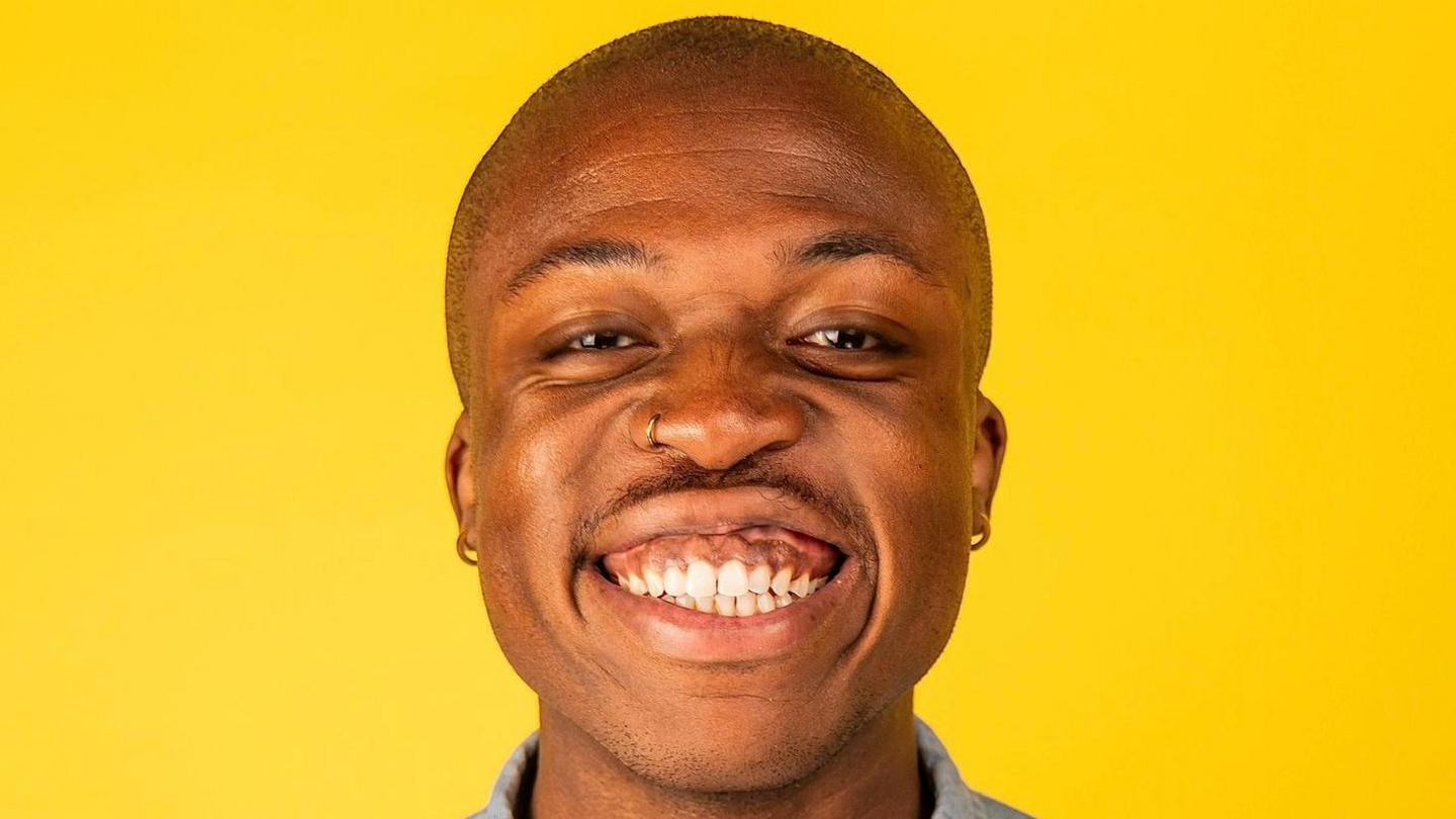 Benjy is bald and is smiling at the camera. He has a gold nose ring and hoop earrings. He is standing in front of a bright yellow background. 