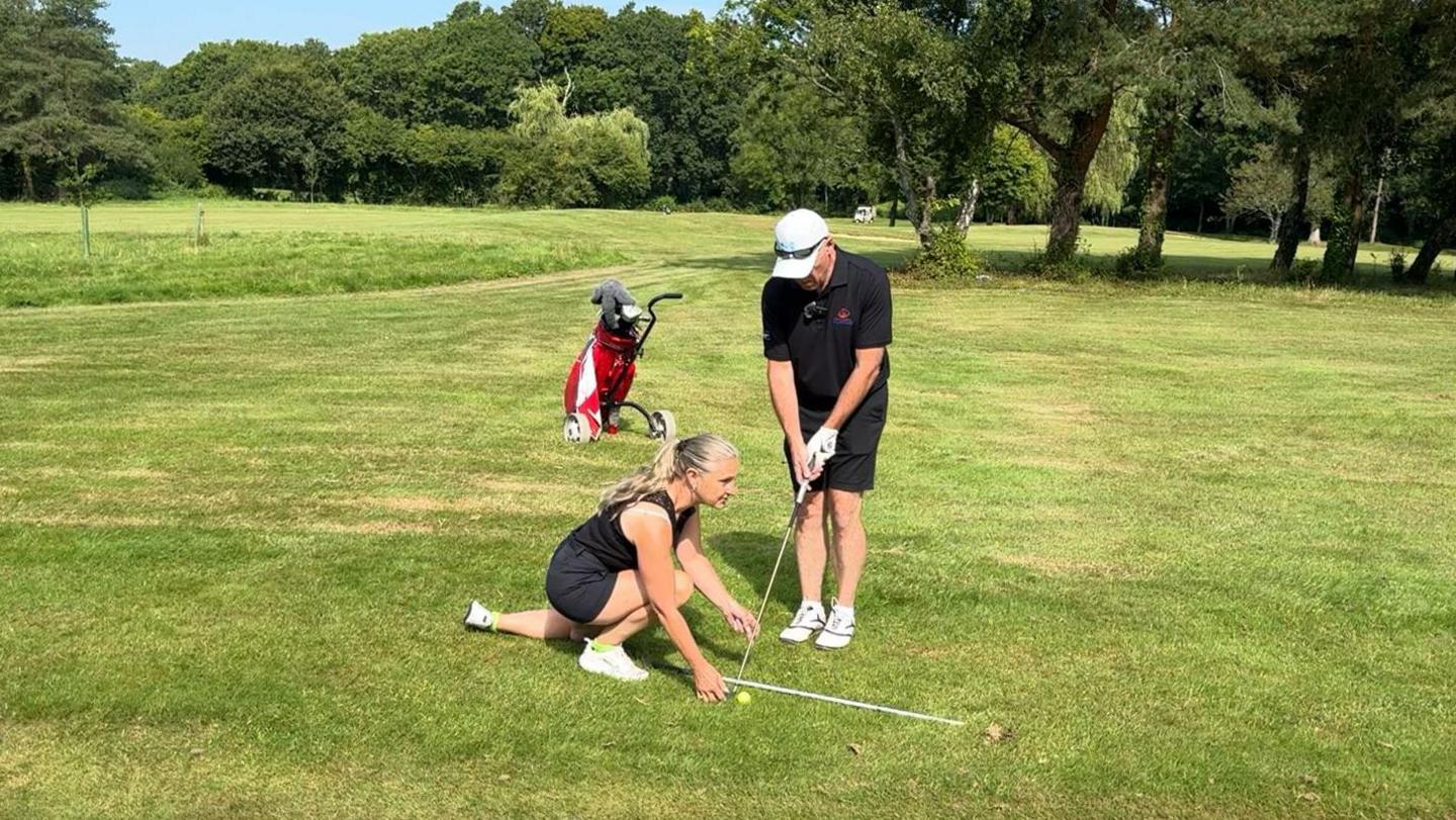 Jane is assisting Simon with his line, she is on the floor with a white stick establishing the line and Simon is stood over the ball preparing for his swing at a yellow golf ball. They are on a lush green course and there is a red and white golf bag just behind them.