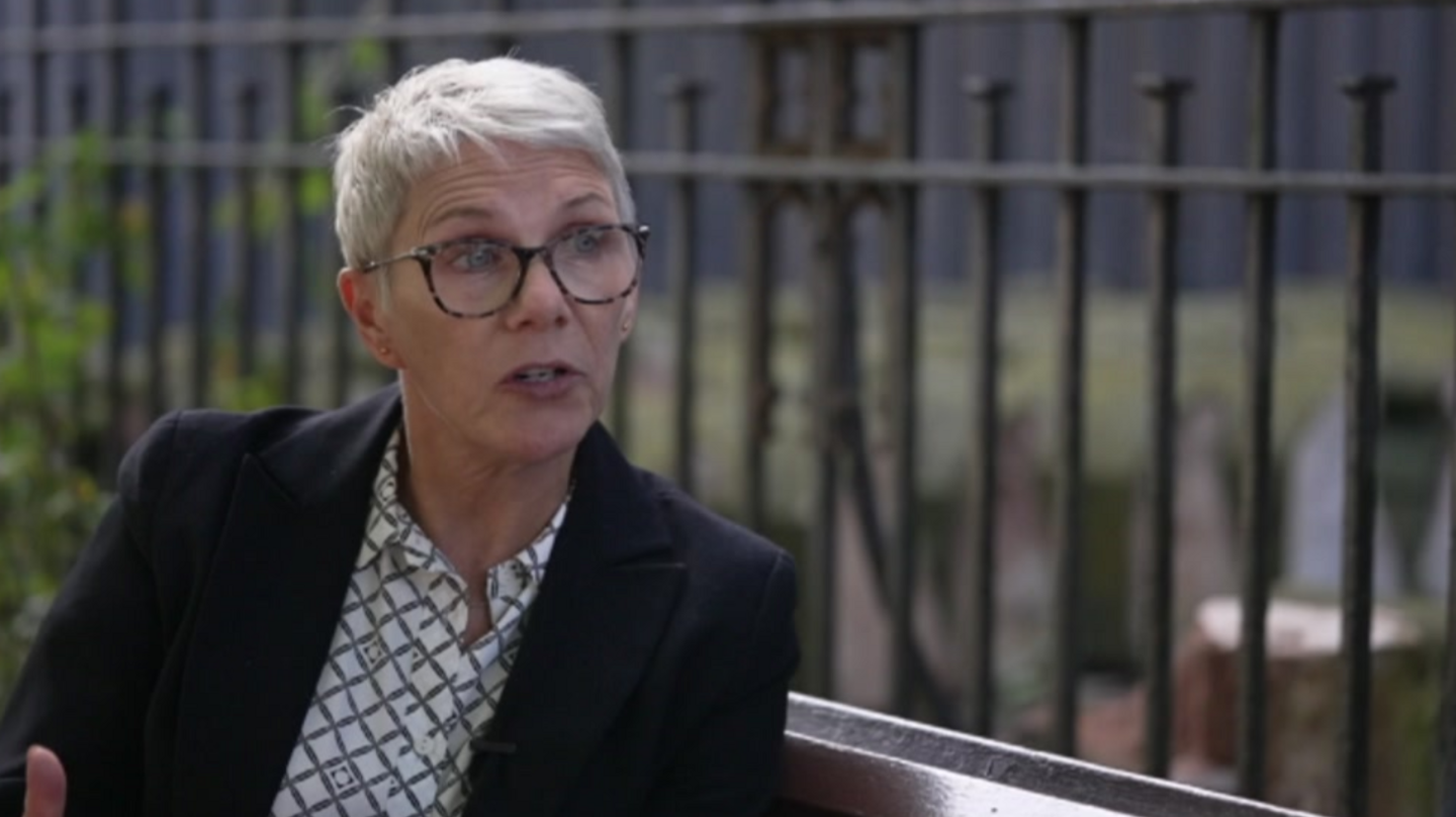 Alison Hinds wearing a dark jacked and white chequed blouse with dark glasses, sitting in front of a fence