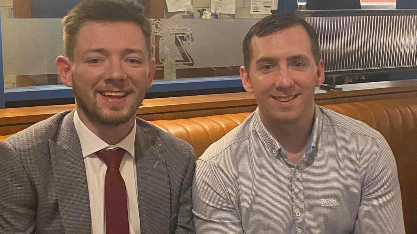 Cahir, left, wearing grey suit and red tie, sits beside Paul, clean shaven with short dark hair, wearing a light coloured Hugo Boss shirt