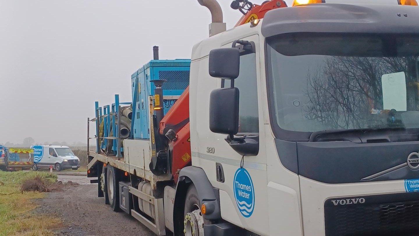 A large Thames Water tanker truck with coiled pipework and big blue containers on the back. In the background there are more Thames Water vans and equipment. It is a grey and misty day.