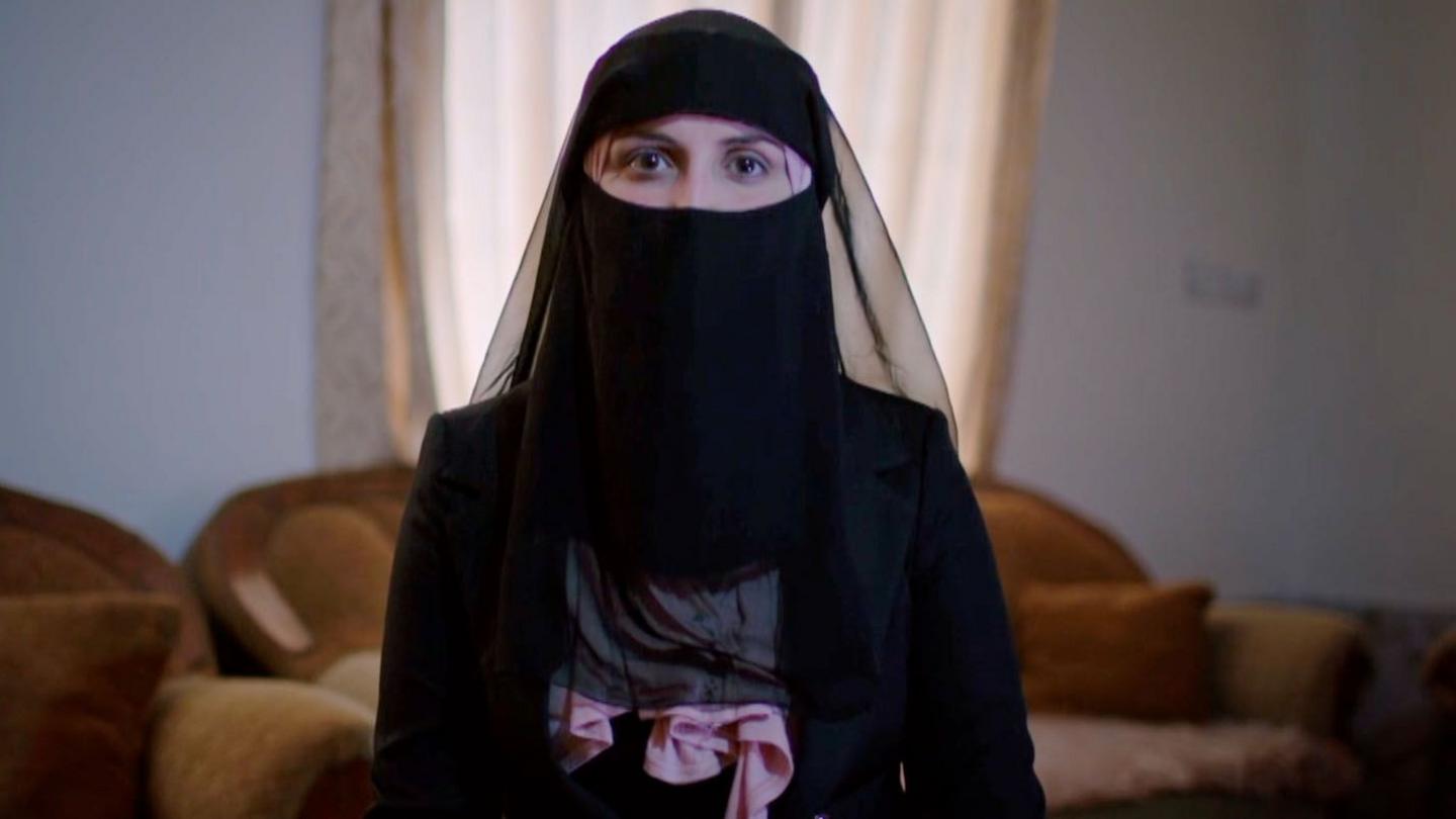 A young woman is seated looking straight at the camera. She is wearing a black robe, a black headscarf and a black veil that covers most of her face, leaving just her eyes visible. A bit of a pale pink scarf can be seen under the veil.