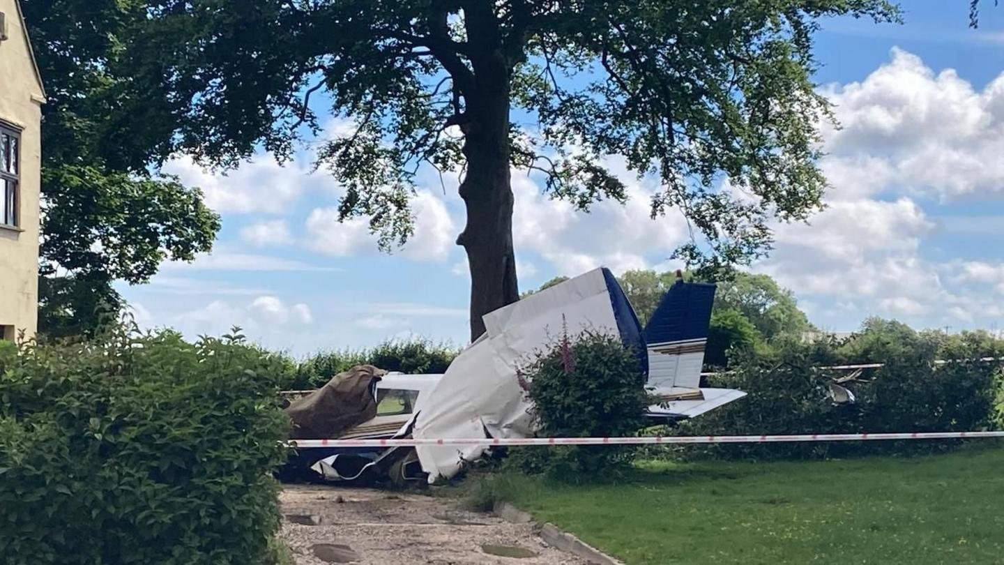 A closeup shot of a partial view of a plane wreckage on a lawn and patio path surrounded by a cordon tape. On the left is a slight view of a house. Behind is a big tree and in front is a bush.