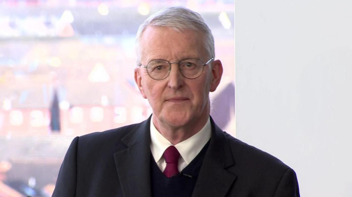 A grey haired man wears circular glasses, as well as a dark suit jacket, a dark jumper, a white collared shirt and a red tie. He is speaking from a podium, and behind him are blurred rooftops of nearby buildings.