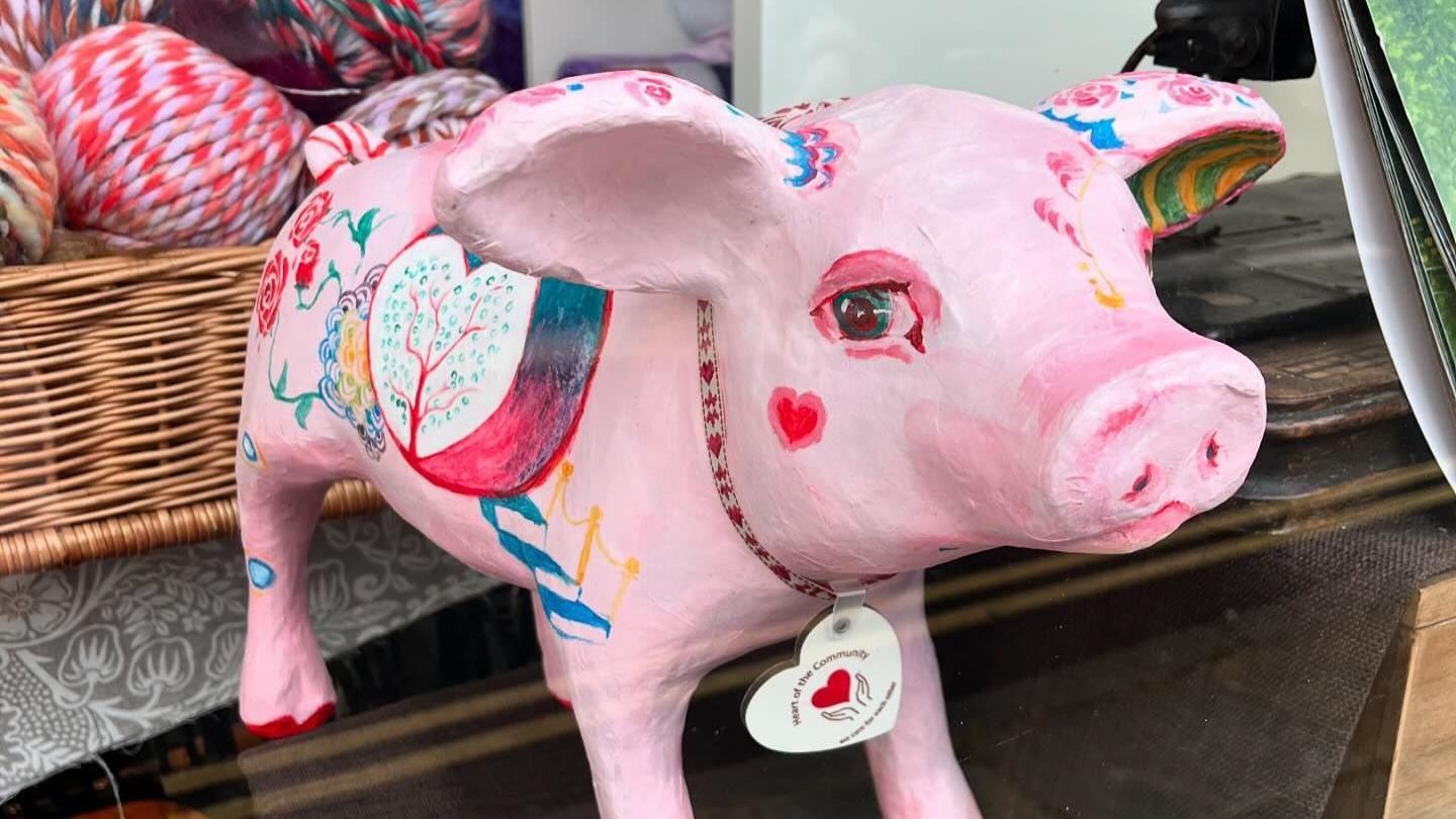 A mostly pink pig sculpture, but with more intricate painted decoration, particularly floral, with lifelike eyes and a ribbon and tag round it's neck.