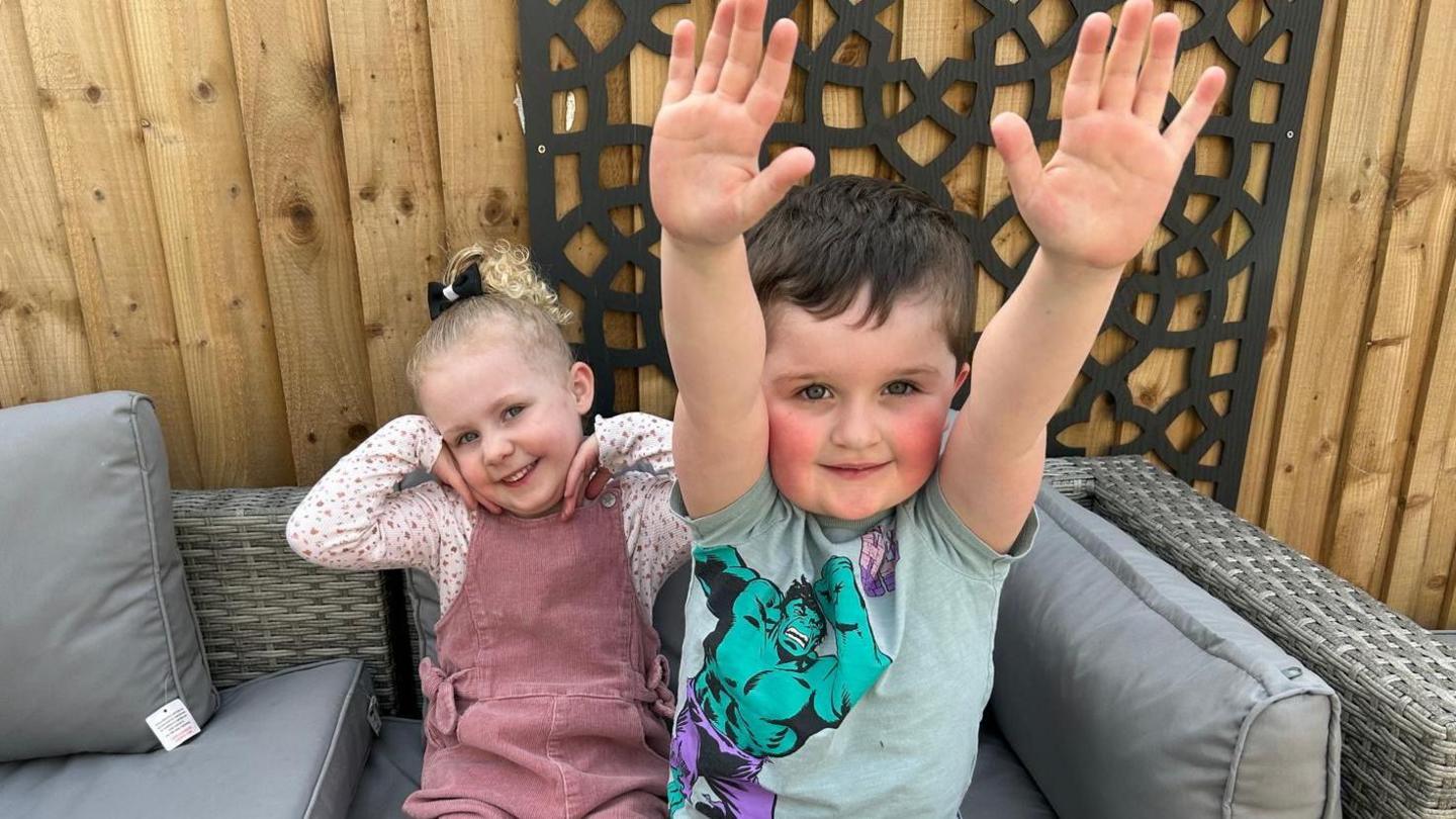 A young girl with blonde curly hair, has her hands to her face smiling in a pose as she sits on a garden sofa, next to her is a young boy with his arms raised in the air, smiling towards the camera, with short brown hair.