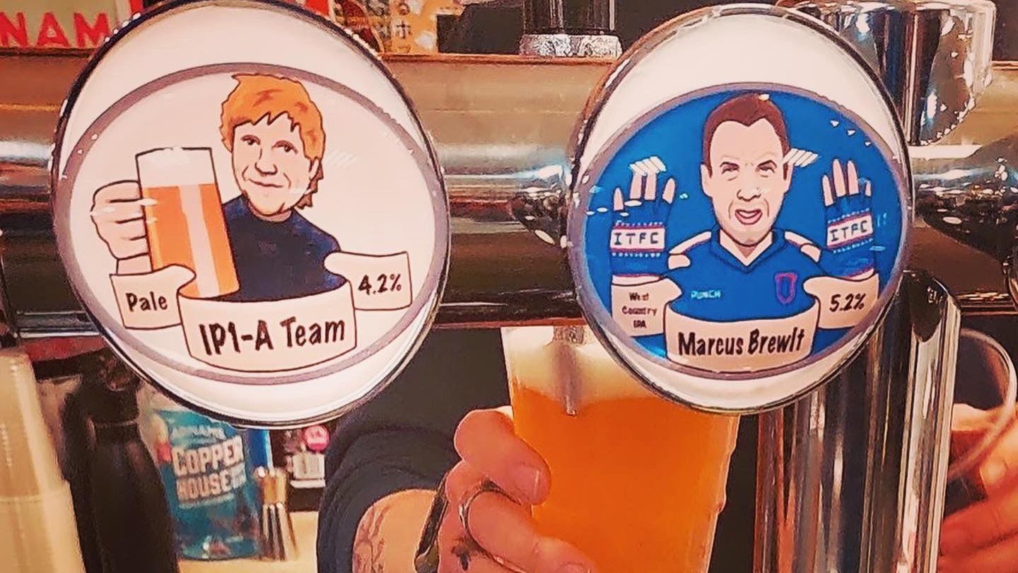 A view of several beers on draught at a pub. An Ipswich Town-themed beer and an Ed Sheeran-themed beer are on display on the front of the taps. A member of bar staff is seen behind the taps pulling a pint.