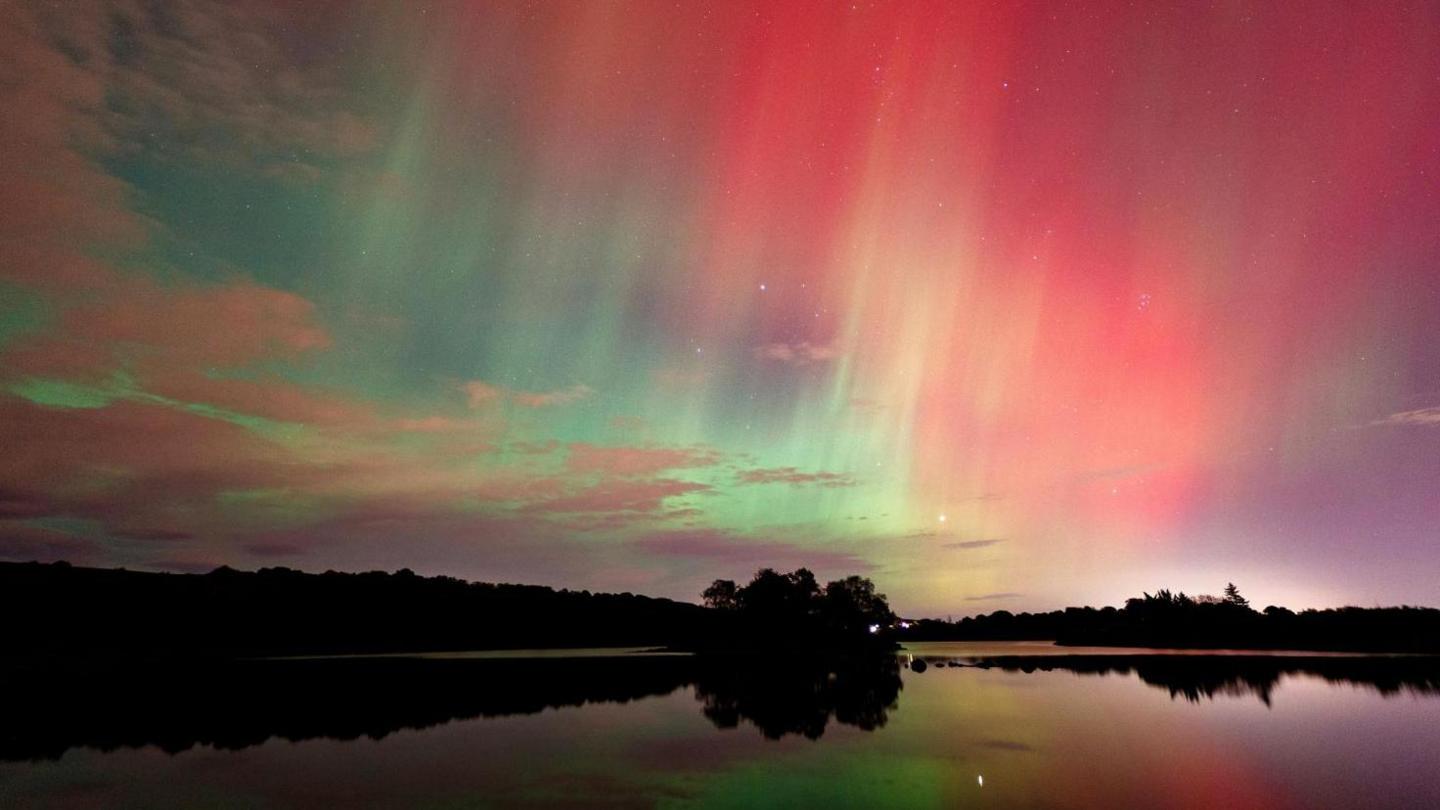 Shimmering green, red and orange lights of the aurora reflected in water and above trees.