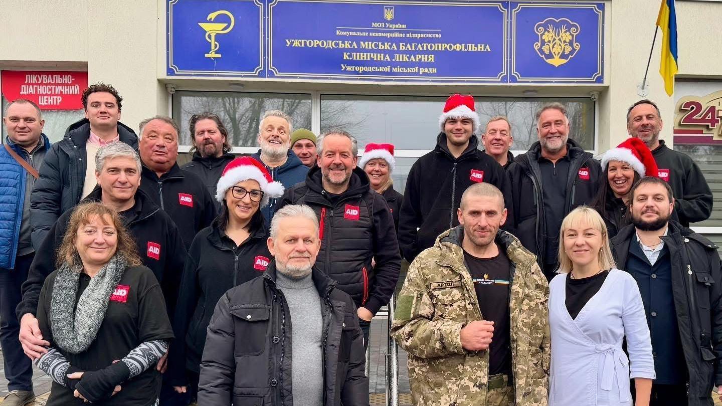 About 20 people stand outside a white building with a blue and yellow sign. They are mostly wearing black coats and red Santa hats. One man is wearing camo army gear and the woman next to him is wearing a purple cardigan.