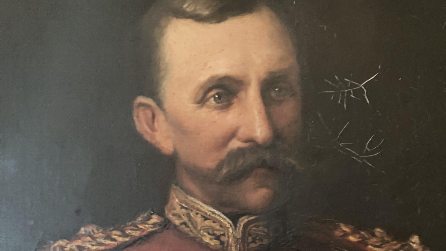 A portrait of Sir William Penn Symons. Sir William is seen wearing a red soldiers tunic. He has a large brown moustache and has been depicted looking off to the right middle distance.