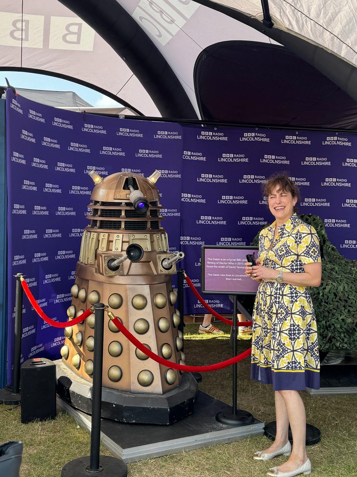 A woman wearing a blue and yellow patterned dress standing next to a gold-coloured model of a Dalek.