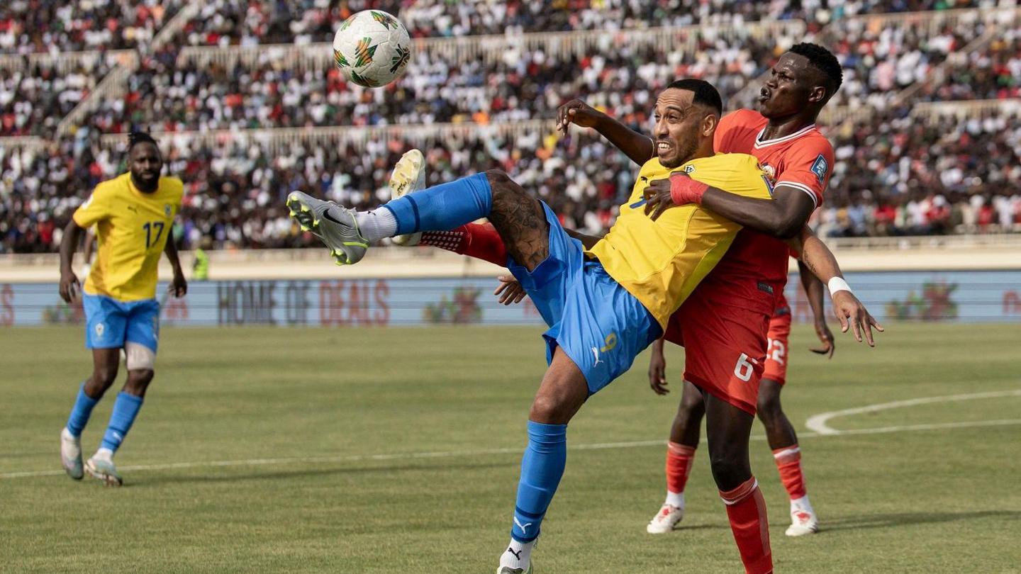 Gabon player Pierre-Emerick Aubameyang, in yellow jersey and blue shorts, is held back by a Kenya player in full red kits as they fight for the ball during a World cup qualifying match in Nairobi