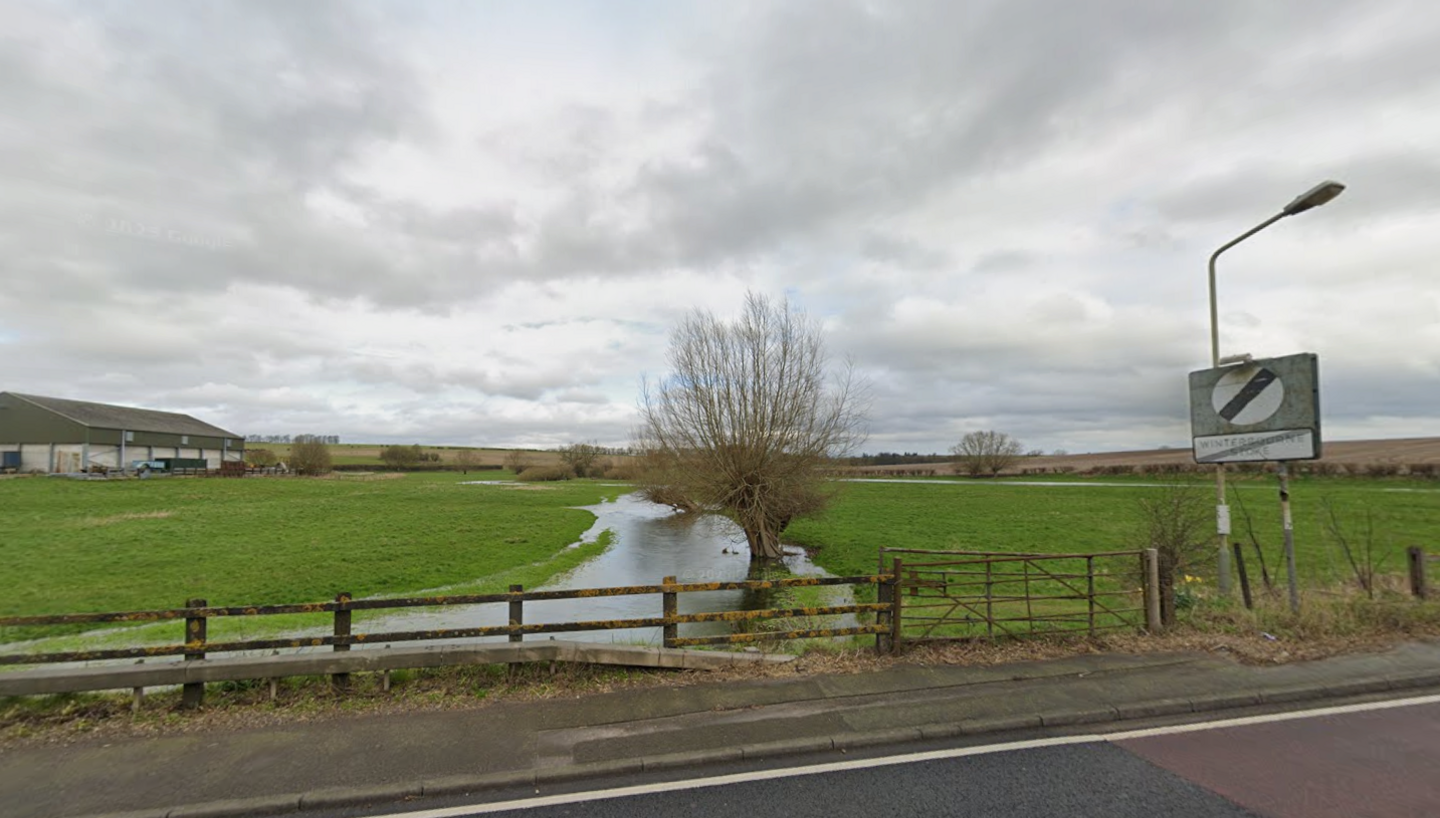Google Maps image of a road with a national speed limit sign and a river surrounded by grass and a farm building in the distance