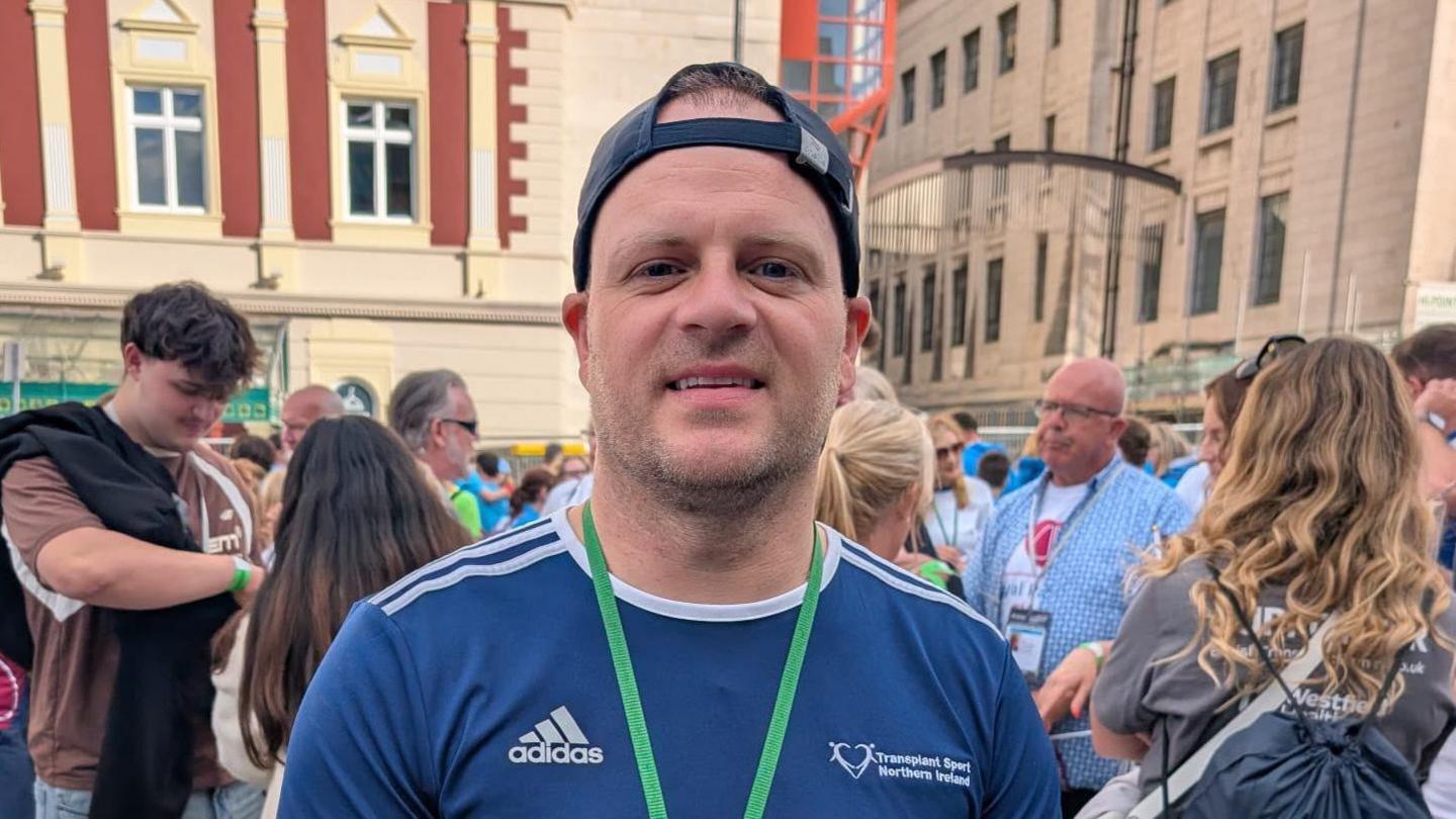 Richie Sheerin is wearing a navy sports T-shirt and a backwards navy cap. He is also wearing a green lanyard. He is standing in front of a large crowd.