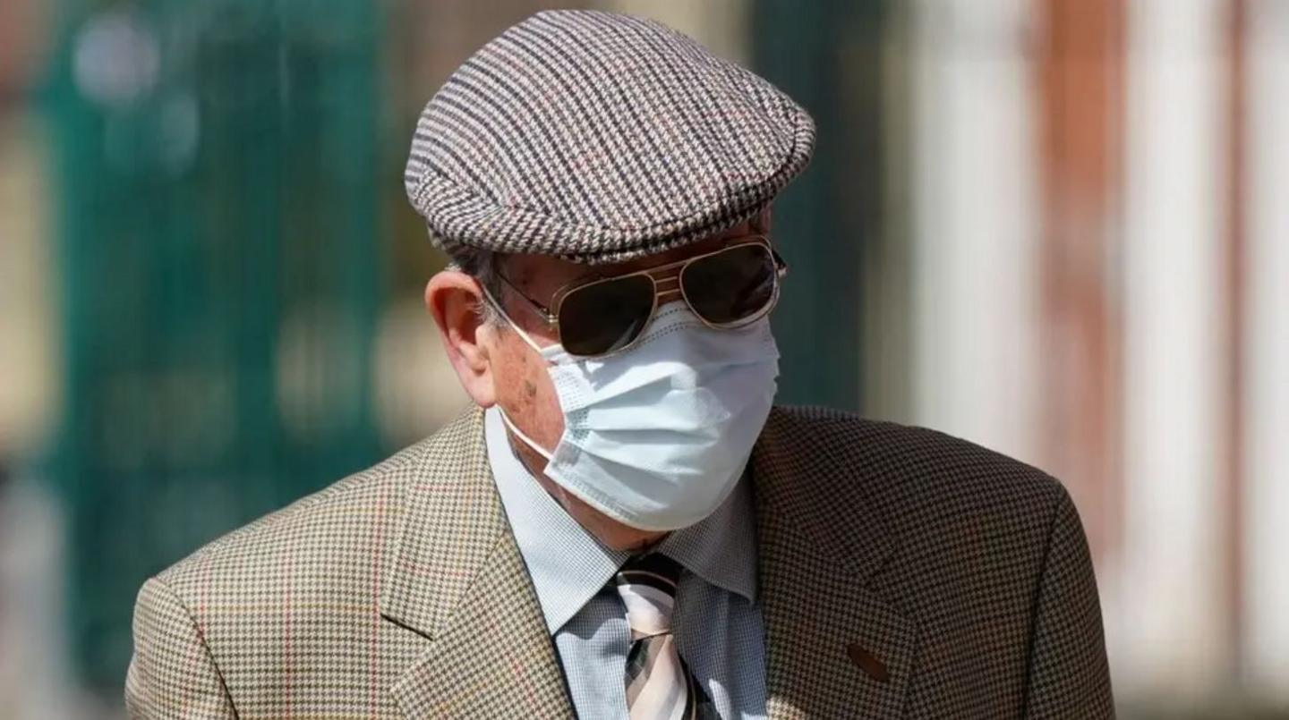 A man in a tweed cap is wearing dark glasses and a mask, which covers much of his face. He also has on a tweed jacket, a grey shirt and a striped tie.