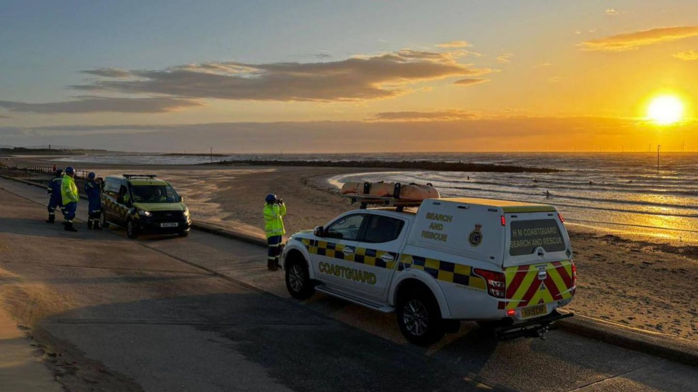 Two rescue vehicles parked along the shoreline with a four men dressed in high visibility clothing with one looking out to see through binoculars. It is late evening and the sun is setting over the sea.