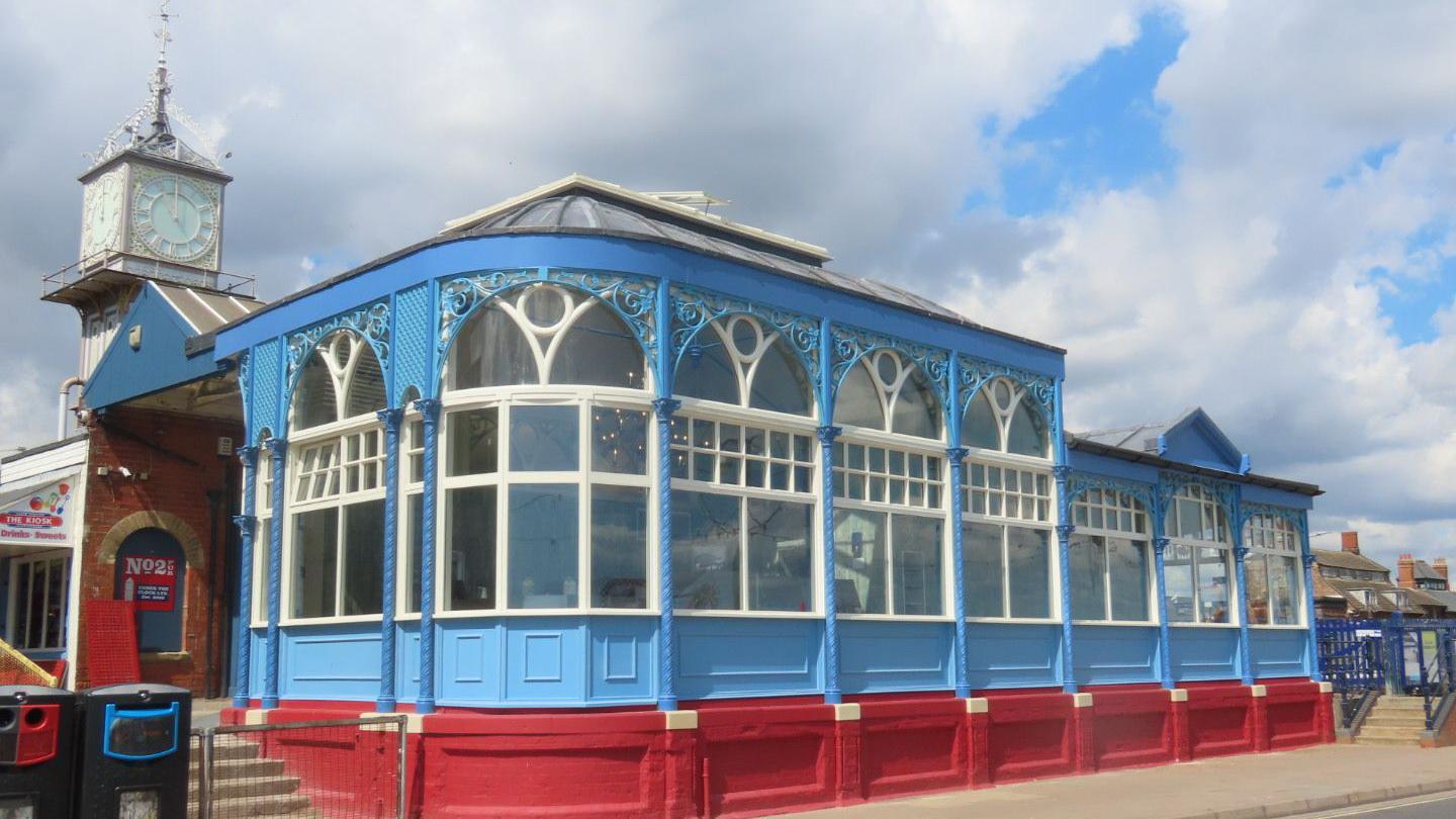 Blue fronted wooden cafe with intricate ironwork decorative framework. The bottom of the cafe is painted red. There are 2 bins standing to the left of the cafe which is near a road and there are steps either side. Behind the cafe building is an ornate Victorian clock tower.