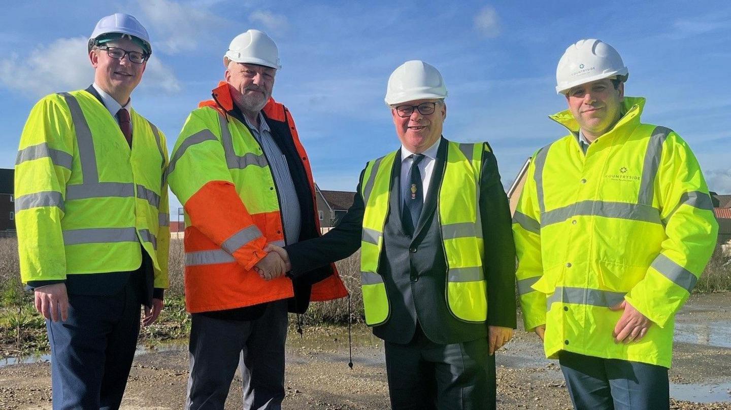 Four men standing on a muddy piece of land wearing hi-vis jackets and hard hats. One of the men is Mark Francois, who is wearing a suit under his yellow hi-vis jacket and is shaking the hand of one of the other men.