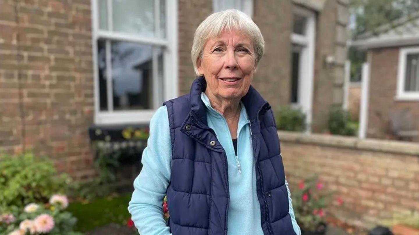 A woman wearing a blue gilet is looking into the camera and standing in front of the window of a house. She has short grey hair and is wearing a turquoise quarter-zip long-sleeve top.