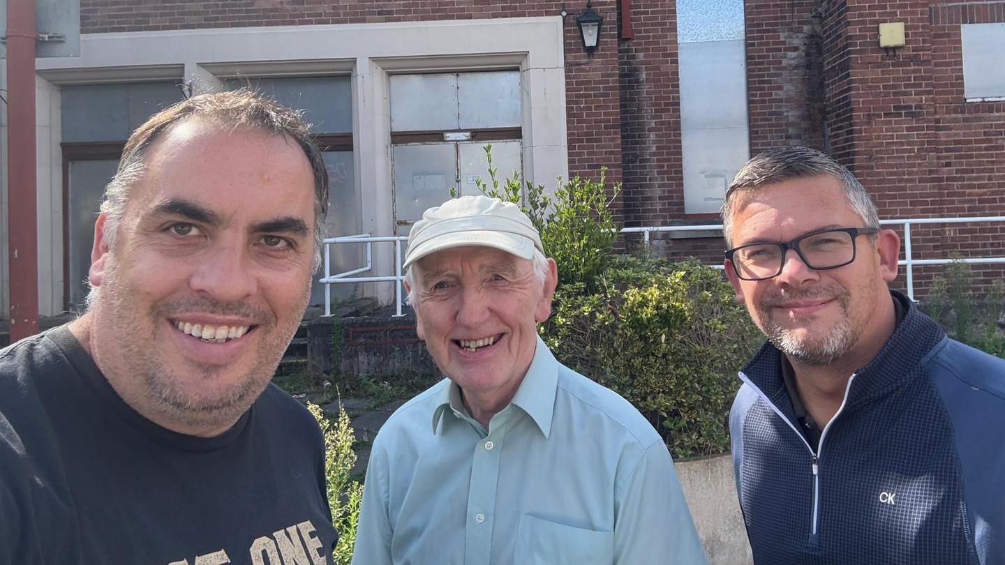 James Bellis (left), Brian Silvester (centre) and Andrew Chandler (right) pose for a selife outside the Flag Lane Baths. The red brick building is boarded up with metal sheets and there are hedges growing along some white railings in front of the building.
