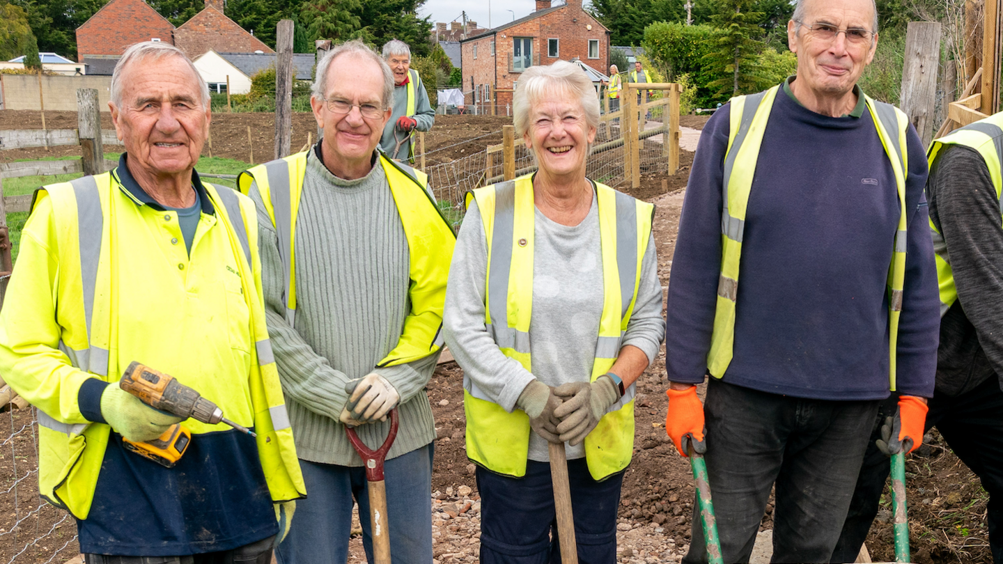Four volunteers standing outside, wearing high-vis jackets and holding spades and a drill. 