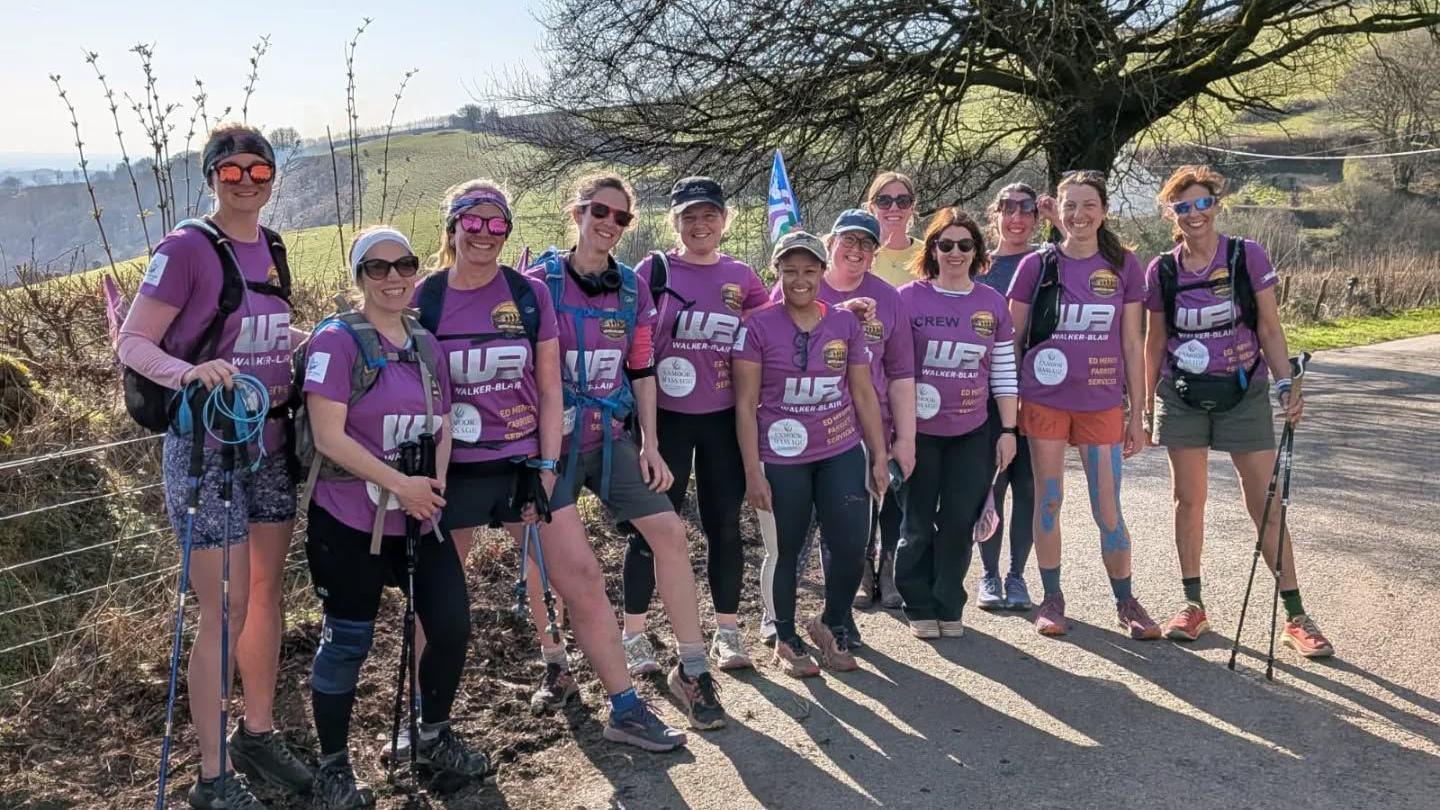 A group of women wearing purple tops, dressed in hiking gear and carrying hiking poles. They are all smiling at the camera, and are photographed in a rural hilly setting