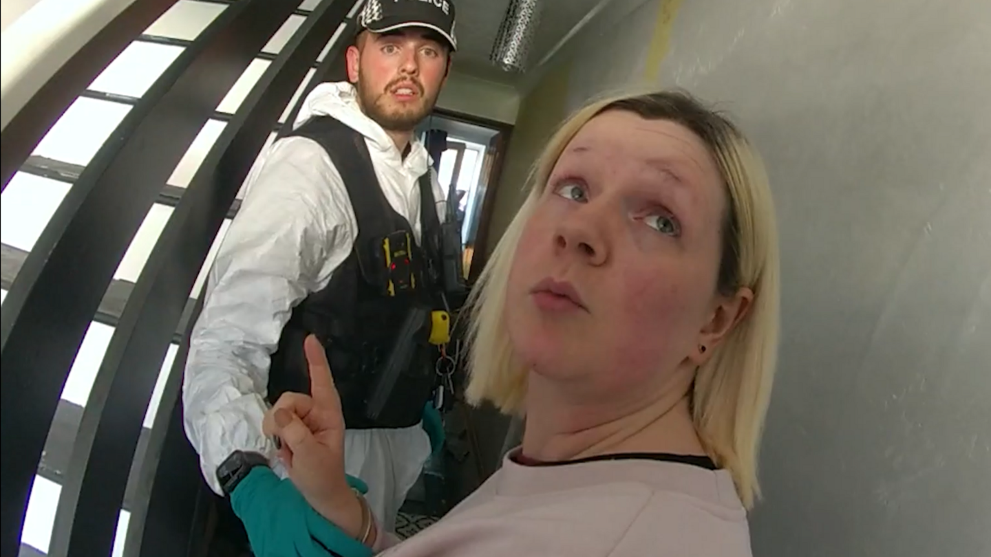 A picture from a body-worn video captured by a police officer. Virginia McCullough is looking over her left shoulder at the officer with a camera. She is wearing a faded pink sweater and being held by a second police officer wearing green gloves, a white forensic suit and black body armour. 