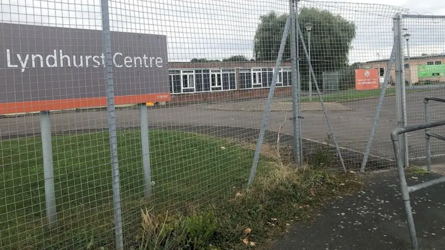 The grounds of Lyndhurst Centre. There is a patch of green grass and a large sign showing 'Lyndhurst Centre'. There is a large metal fence and tarmac on the ground. 