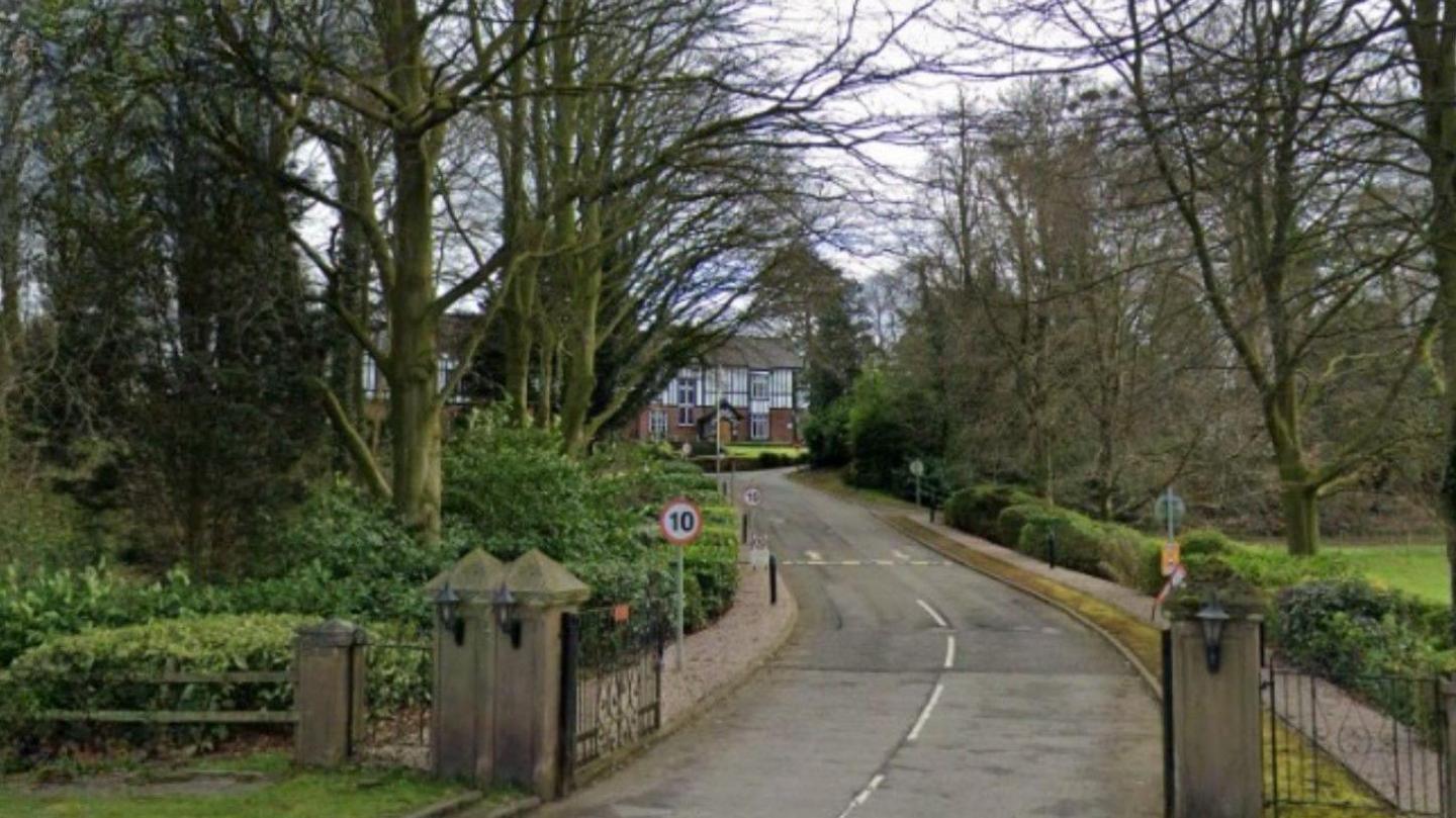 A building, white and black on the exterior of the first floor, is in the distance. Closer to the camera, there are trees on both sides of the road and  a gate leading to the road is open, near a 10mph sign.