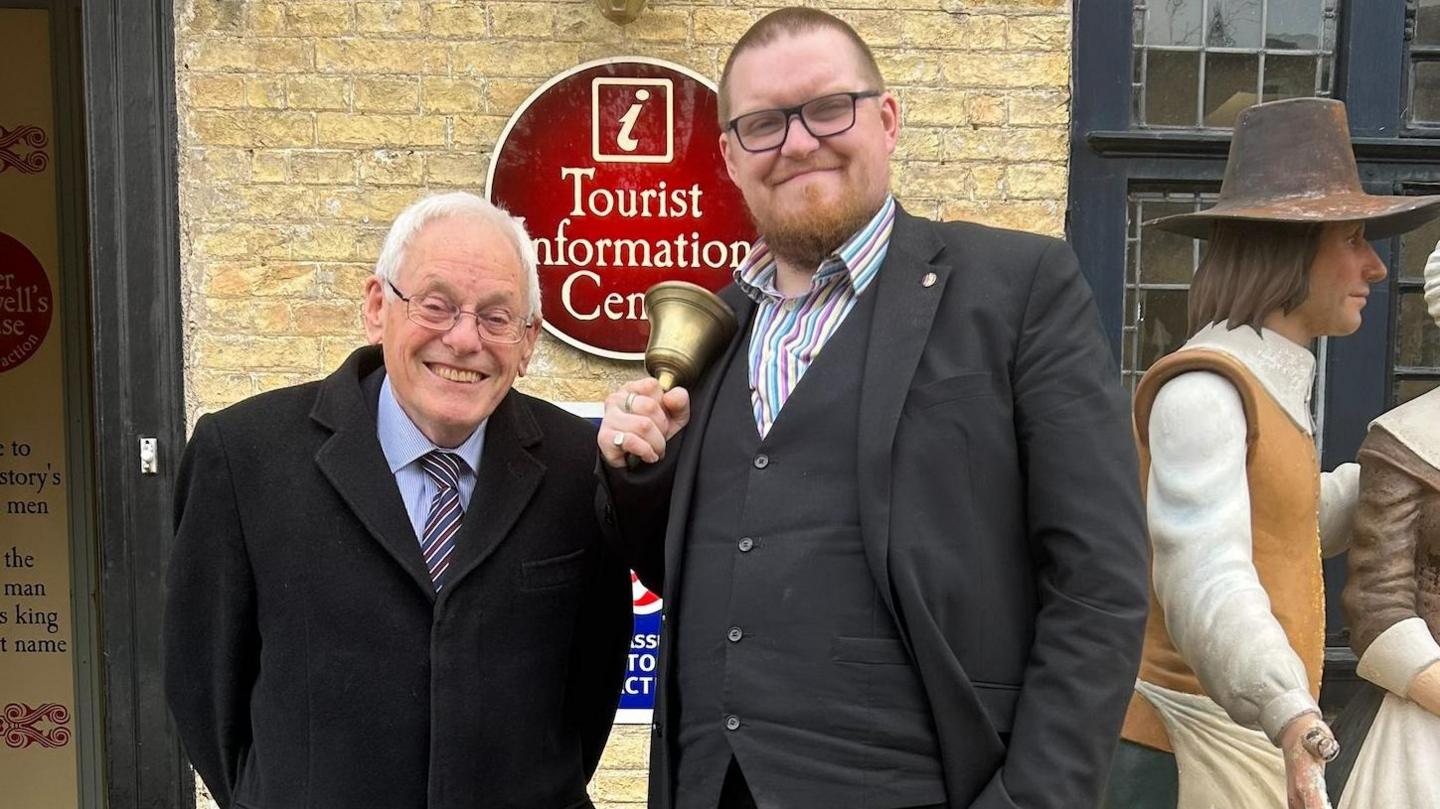 Chris Phillips, City of Ely Mayor, is standing next to Matthew Routledge, the new town crier. Chris is wearing a black coat with a blue shirt and a striped tie. Matthew is wearing a black suit and a striped shirt. He is holding a gold bell. They are standing outside the Tourist Information Centre and Cromwell Museum