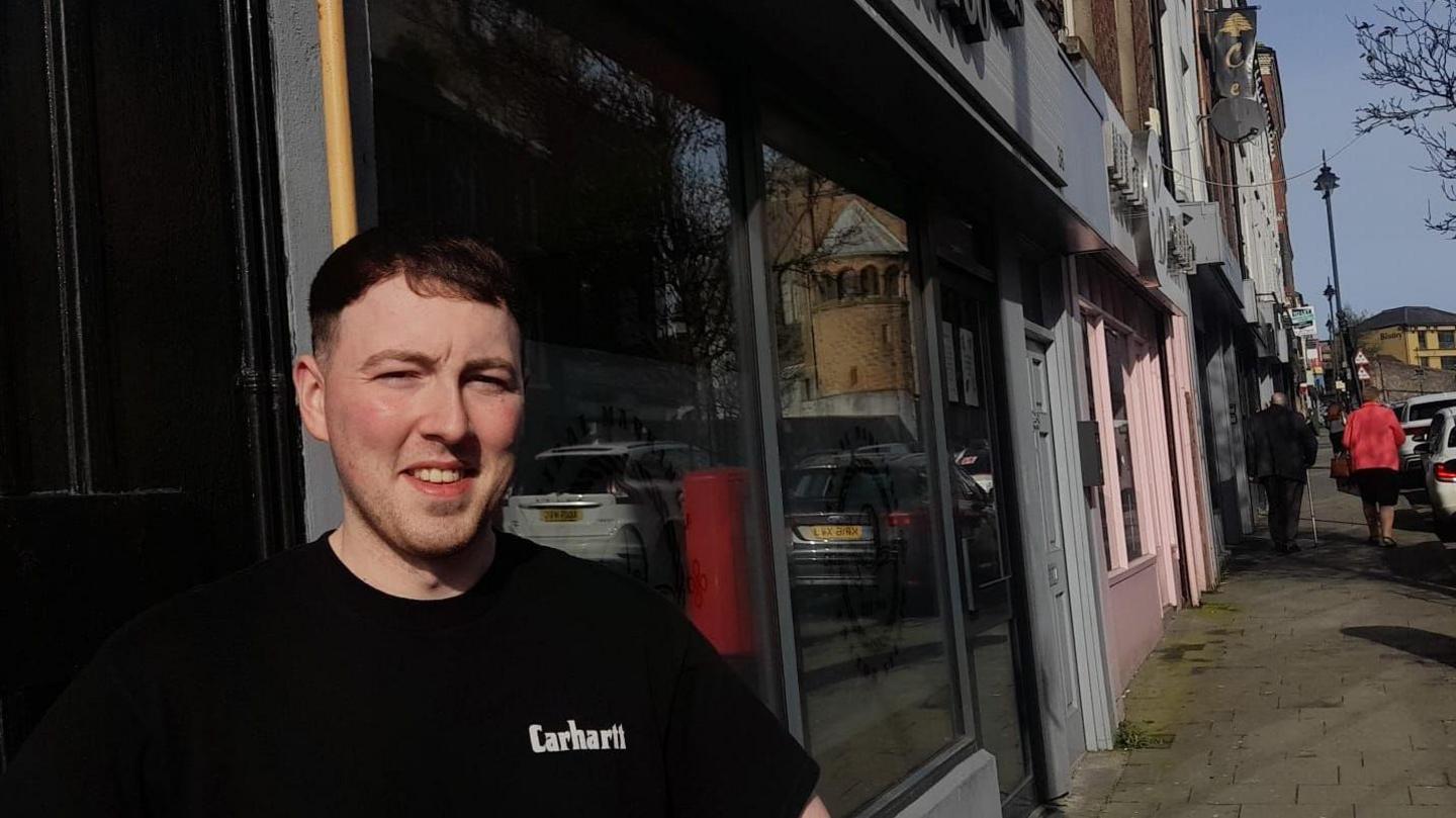 A man stands outside a row of glass fronted shops , two people walk past to his right. The man is wearing a black tshirt, and is squinting to block the glare of the sun