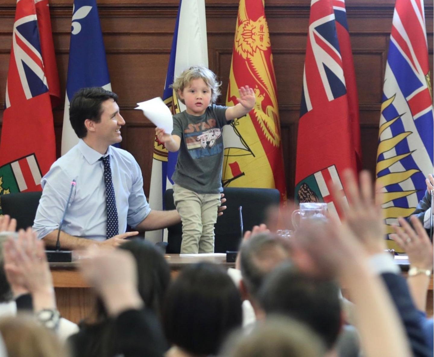 Trudeau junior stands on his chair in a press conference briefing room, holding up his hands to calm the press, who can be seen in soft focus with hands raised in the foreground.