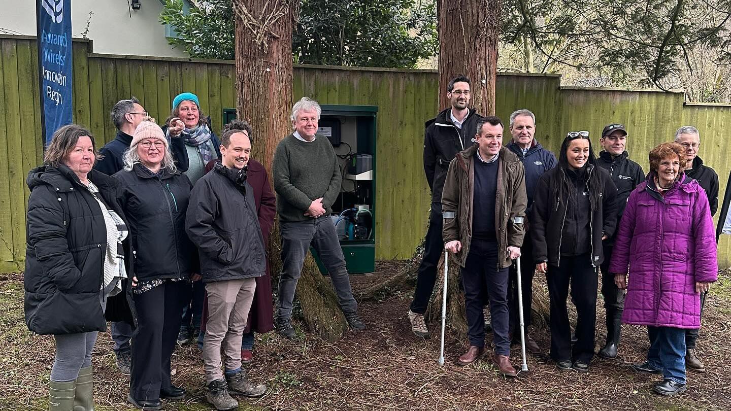 A group of men and women are standing next to a green wooden fence which has a dark green metal cabinet attached to it. The cabinet contains the monitoring equipment. 