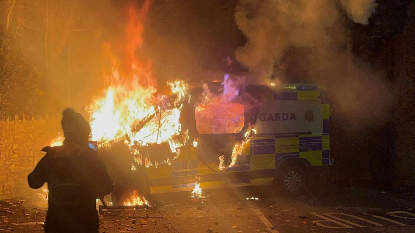 A police van is on fire. There is a person standing in front of it, they have a hat on. The front half of the van is almost entirely burnt. The background is a blur of flames and smoke. There is a stone wall behind the van. 