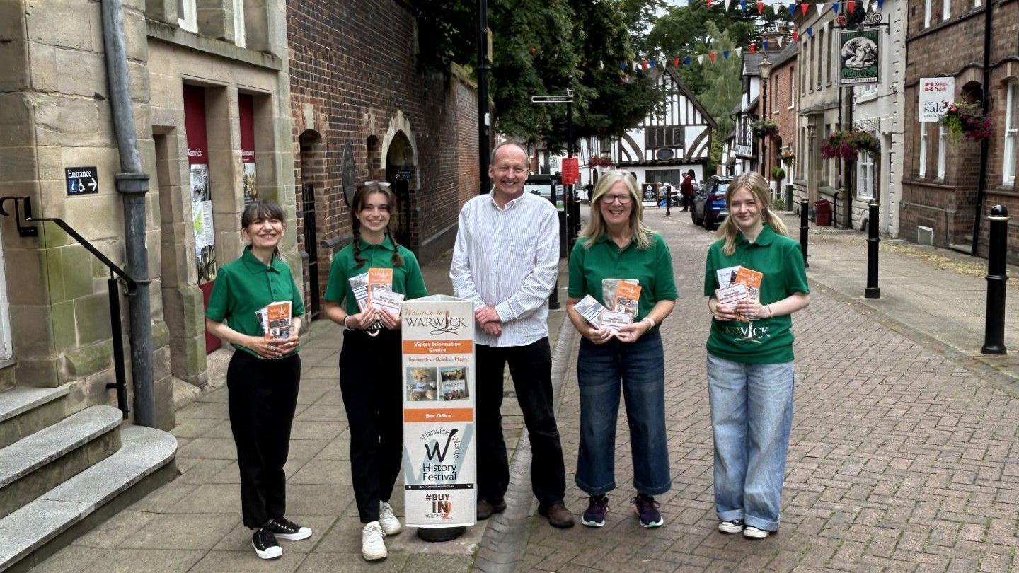 Four women in green T-shirts stand on a street holding leaflets. in the middle of them stands a man in a white shirt. In front of him is a sign advertising Warwick.