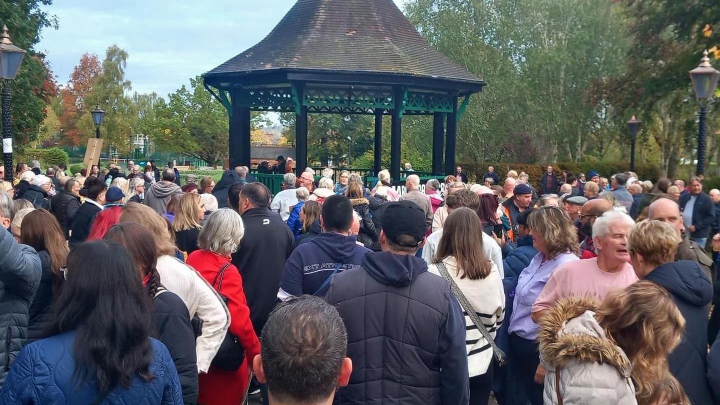 A crowd of dozens of people gather in front of a bandstand in a large park area. Many are wearing coats and we can see the backs of their heads. The bandstand has a black tiled roof with signs of green moos and several pillars supporting it from the ground. Behind it are several tall trees.