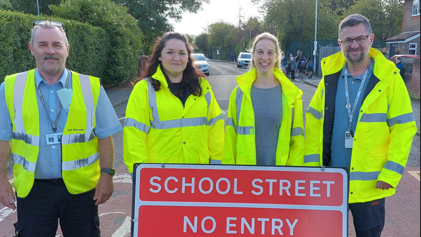 Two women and two men standing on a residential road wearing high-vis yellow jackets. One of the men is wearing a whistle around his neck. In front of them is a red and white sign which reads "school street no entry". There are some young children wearing backpacks in the background.