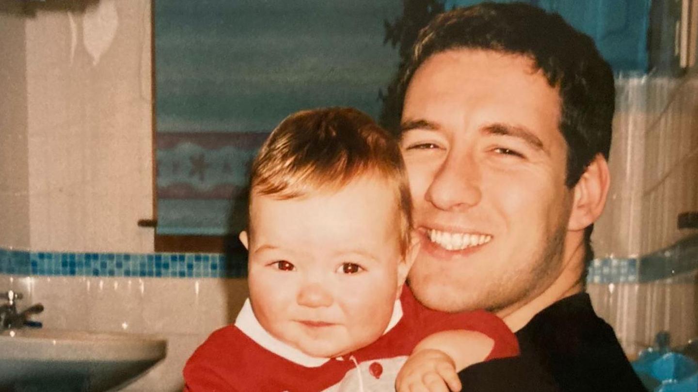 Alistair is pictured with one of his young sons in a bathroom. Alistair is smiling.