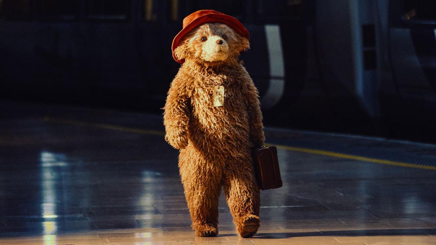A first look at Paddington in the new West End musical. He's wearing his famous red hat, carrying a suitcase and has a note tied around his neck with string. He is dramatically lit as he walks down a railway platform, his brown and gold fur ruffled. He moves freely, without a human directly operating him or puppet strings.